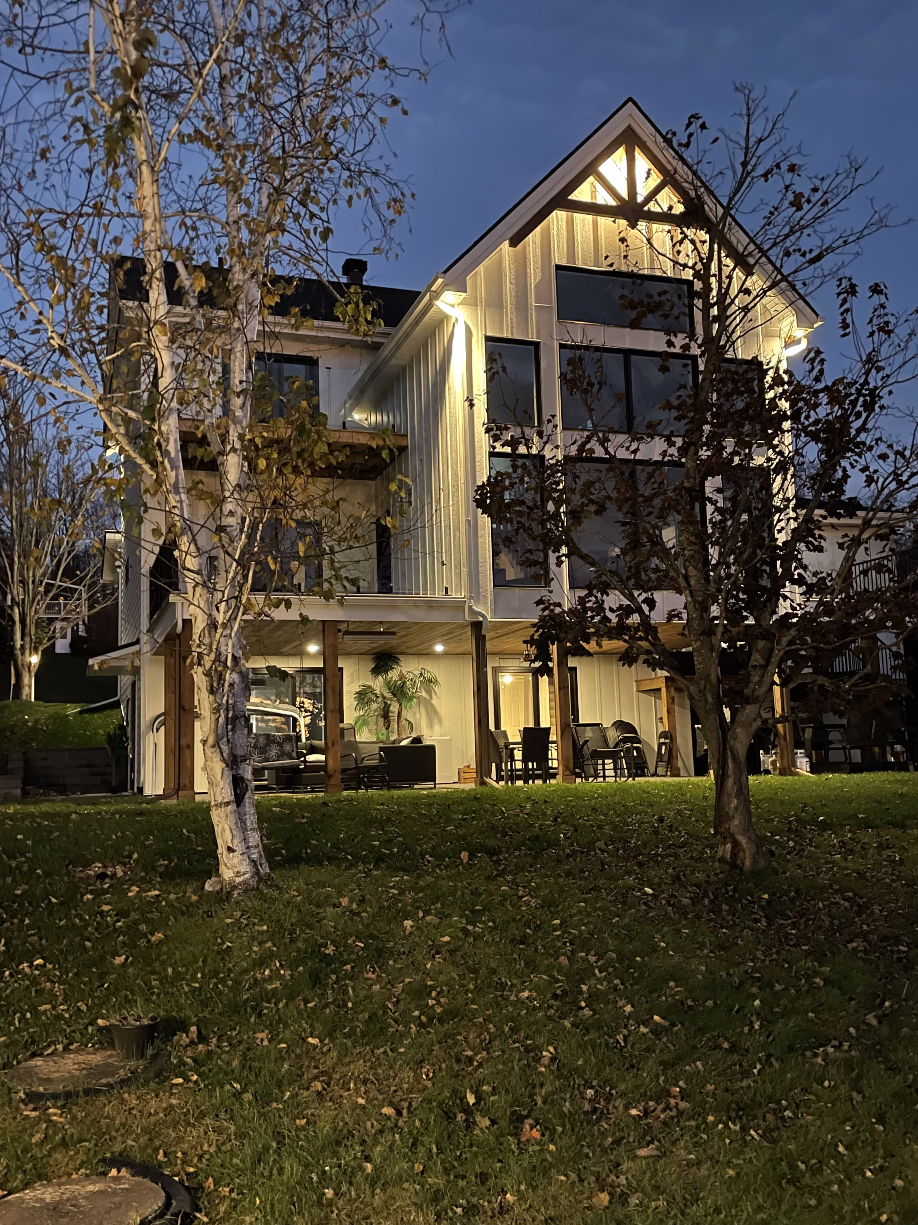 Résidence à pignon illuminé, grandes fenêtres et terrasse, entourée d’arbres au crépuscule